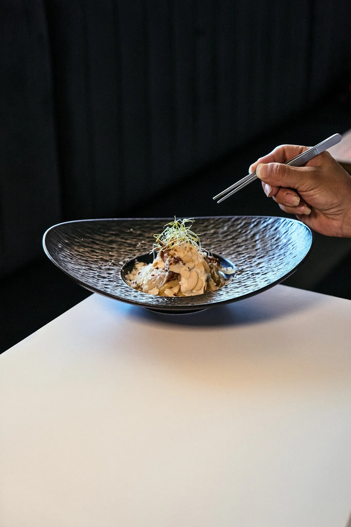 Una mano con palillos sobre un plato negro de cerámica texturizada con un bocado de cocina fusión, en una mesa blanca