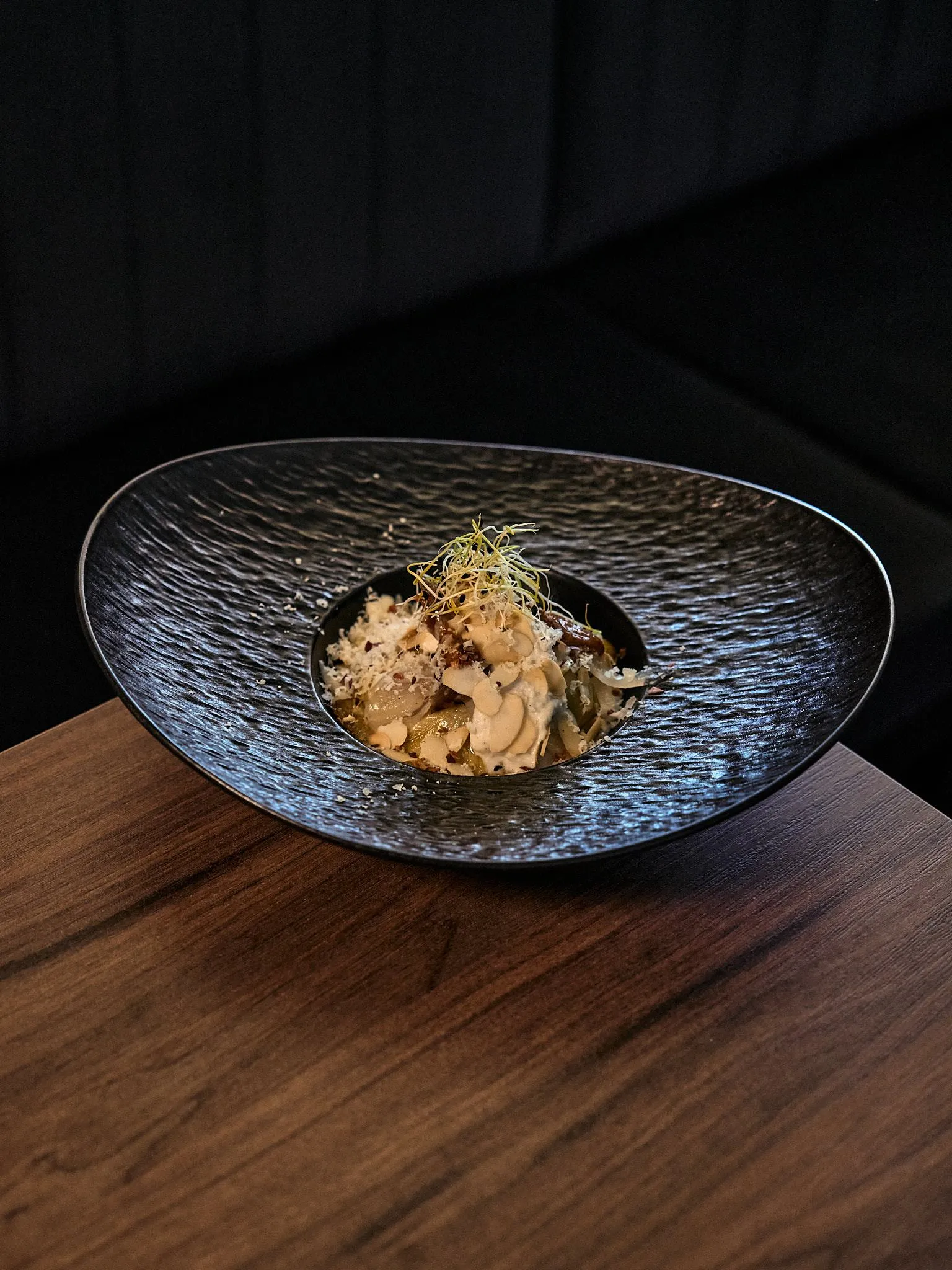 Plato negro de cerámica texturizada con un bocado de cocina fusión sobre una mesa de madera