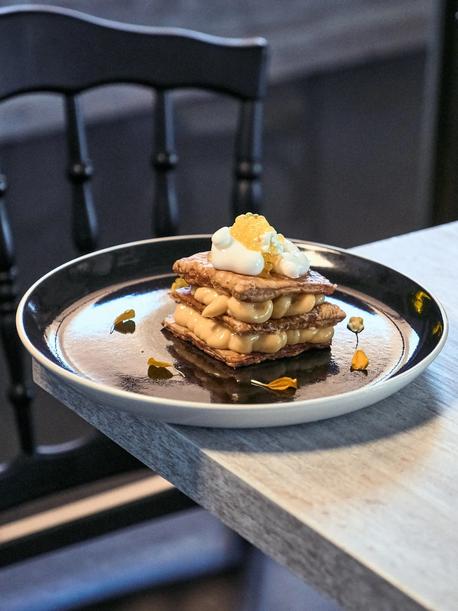 Milhojas de crema con huevas y flores en un plato blanco, junto a una silla negra